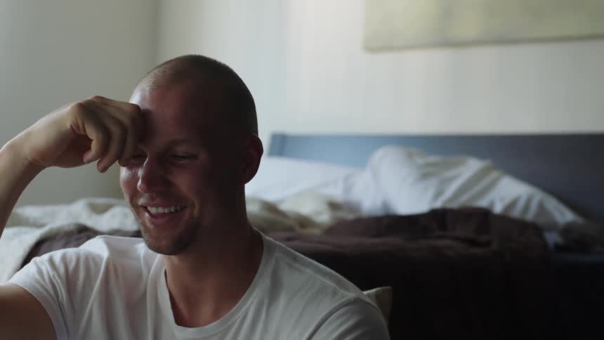 A man sits on the floor leaning on the bed and laughs while looking at photos
