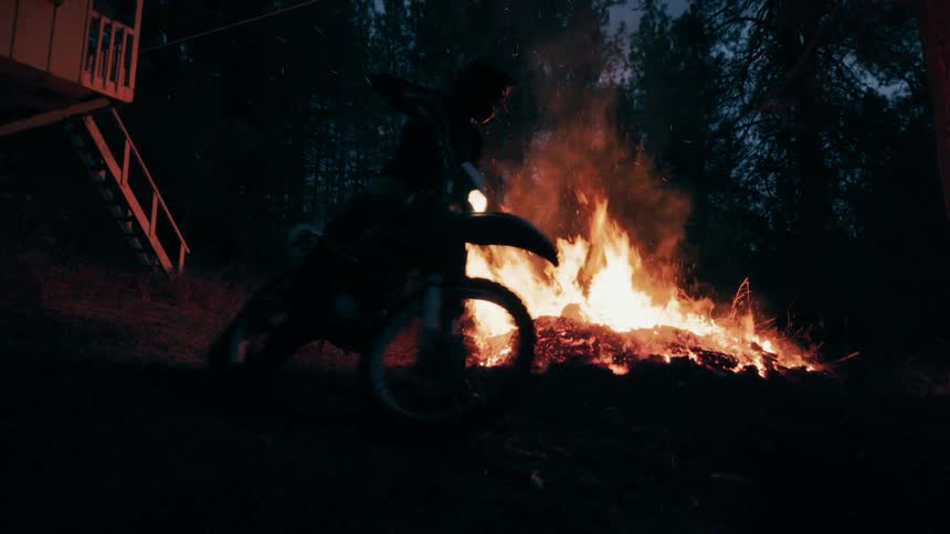 A dirt bike rider speeds past a blazing campfire at night the flames casting dramatic light and sparks against the dark forest backdrop