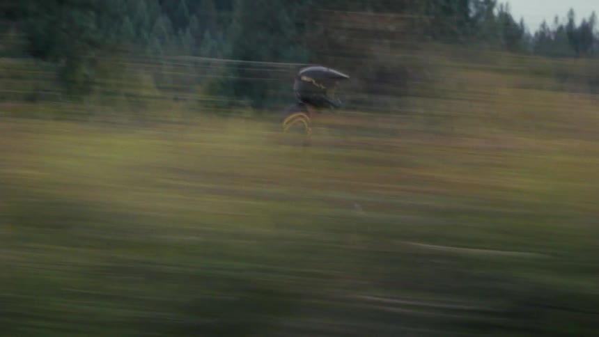 A motorcyclist wearing a black helmet and jersey rides a red and white dirt bike across a grassy open fieldthe camera uses a fast panning motion creating a blurred foreground and background that emphasizes the speed of the rider