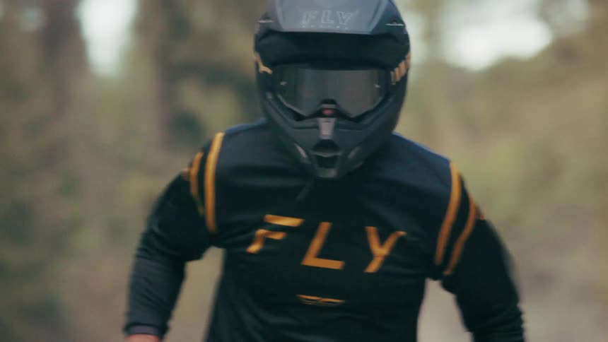 A person wearing a black and gold fly racing jersey and a matching helmet rides a dirt bike down a gravel path the rider navigates through a wooded area as the camera captures them from a frontfacing perspective
