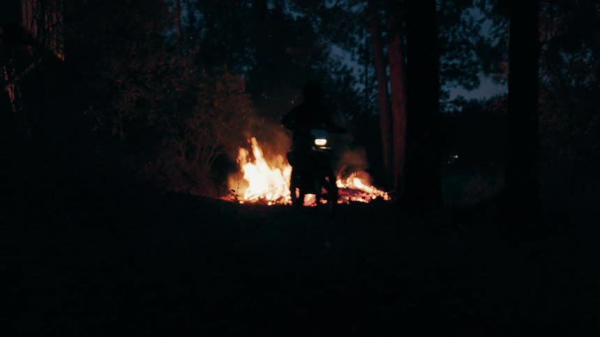 Silhouette of a biker in the dark riding a motorcycle away from a campfire in the woods outdoors during night time