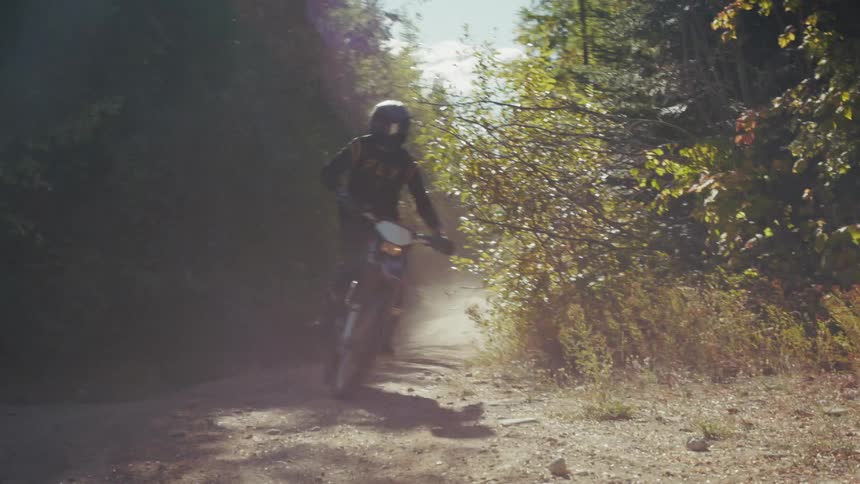 A motorcyclist wearing a black helmet and jersey rides a red and white dirt bike down a dusty sunlit path through a wooded area as the bike passes the camera it kicks up a cloud of dust and reveals the number 86 on the rear side panel