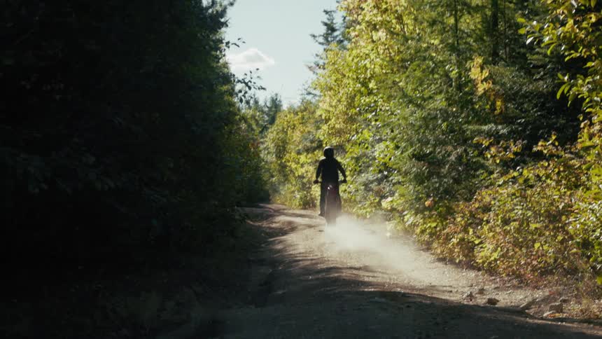 A man riding a red dirt bike speeds away from the camera down a narrow sunlit forest path