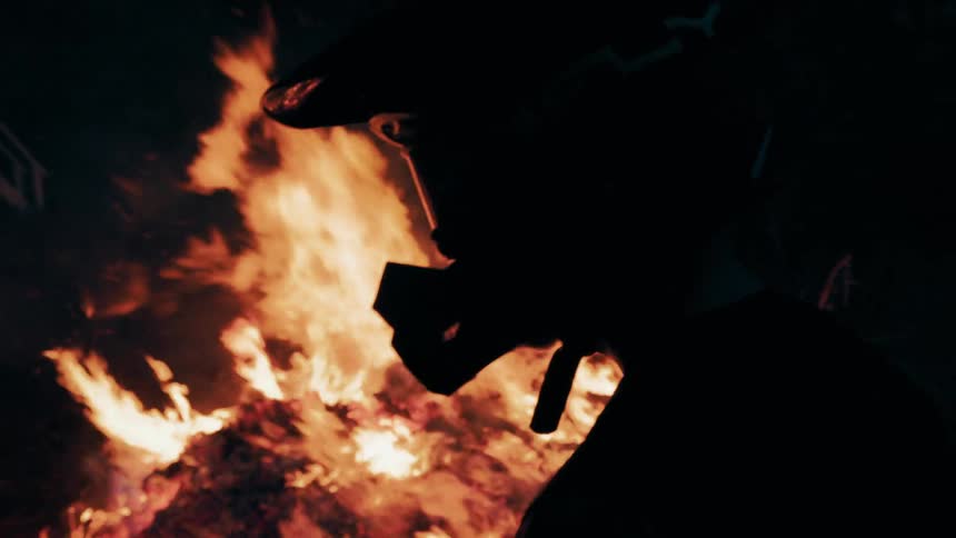 Silhouetted person wearing a mask facing intense flames in a nighttime setting