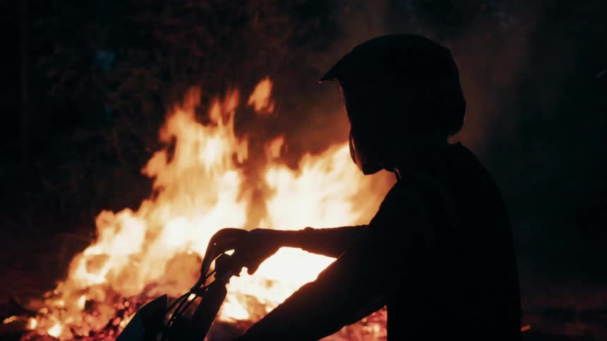 Silhouette of a person wearing a helmet on top of a motorcycle next to a bonfire on a grove outdoors during night time