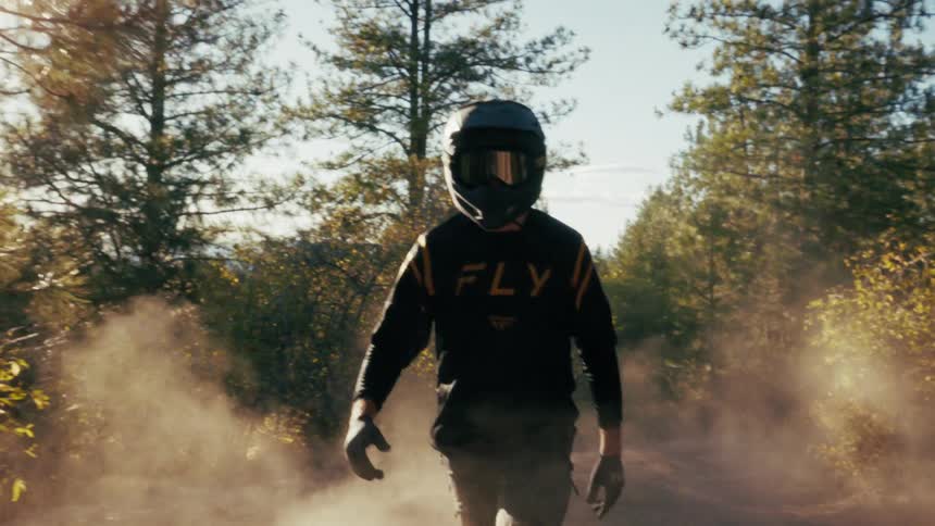 A person wearing a fullface motorcycle helmet and jersey walks toward the camera through a cloud of dustthe scene is set on a wooded trail during the golden hour with sunlight filtering through the surrounding pine trees