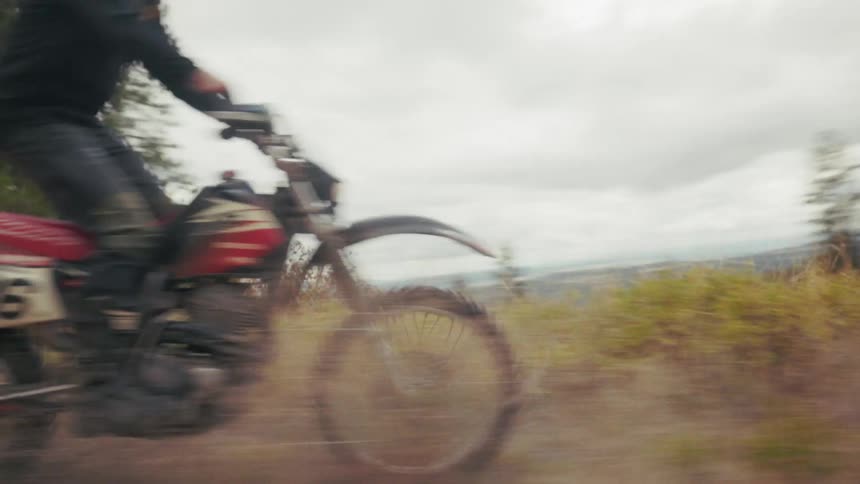A dirt bike rider speeds along a dusty trail kicking up a cloud of earth as they navigate a turnthe camera follows closely from a low angle capturing the surrounding pine trees and autumncolored brush