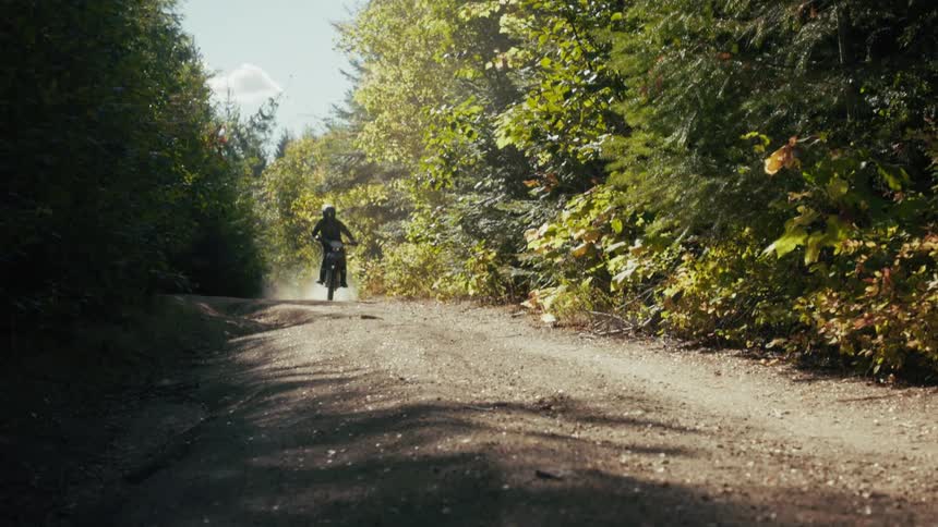A person on a motorcycle speeds down a dirt path through a sunlit forest the rider kicks up a cloud of dust while approaching and passing the camera