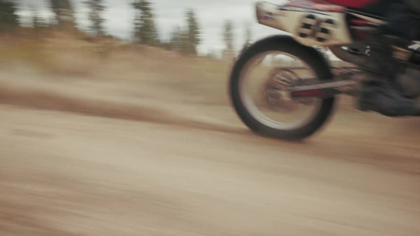 A close up side view shows a dirt bike racing quickly down a dirt path and kicking up dust