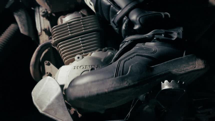 The physical contour of the motorcycles gear shifter it is a rugged metallic component with a serrated or toothed top edge designed for grip extending horizontally at the bottom right of the frame where the riders boot makes contact