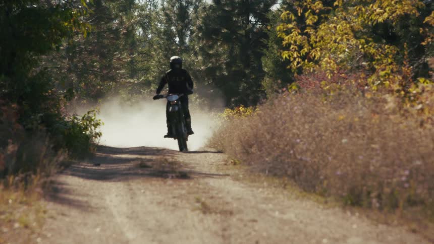 A motorcyclist speeds toward the camera on a dusty forest trail kicking up a thick cloud of dirt