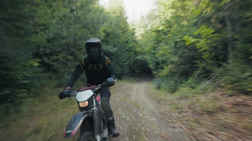 A person wearing a black and yellow jersey and a fullface helmet rides a dirt bike down a narrow forested trailthe camera moves in front of the rider capturing the surrounding dense green trees and the uneven dirt path during the daytime