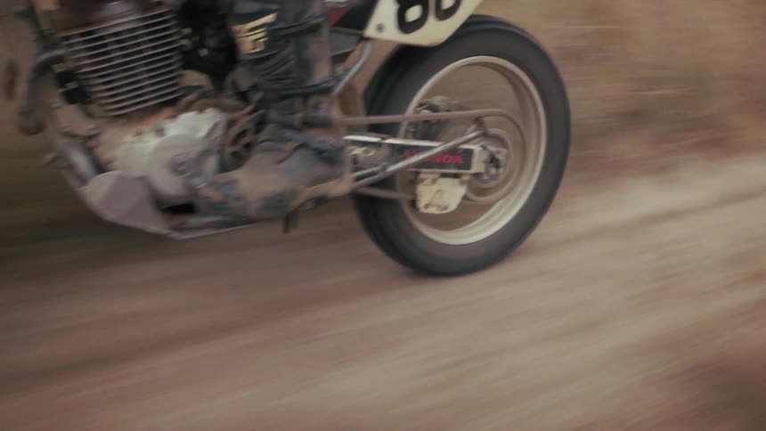 A closeup tracking shot captures a man riding a motorcycle along a dusty dirt road highlighting the mechanical movement of the engine gears and rear tire as the bike speeds through a rural landscape