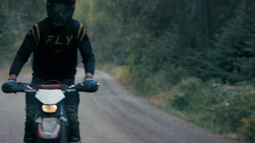 A motorcyclist wearing a black helmet and a black and yellow jersey rides a white dirt bike down a narrow paved road lined with dense treesthe rider approaches the camera while navigating a slight curve in the road kicking up light dust as they pass