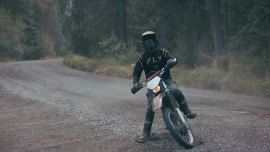 Man wearing a helmet and dark clothes with text on it riding a numbered motorcycle on a road surrounded by a forest outdoors during day time