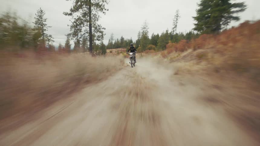 A dirt bike rider in dark gear navigates a winding trail through a hilly forested landscape under an overcast skythe camera follows closely from a low perspective capturing the highspeed motion as the rider kicks up dust while passing through dry b