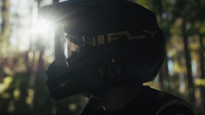 A closeup shot features a person wearing a dark motocross helmet and goggles standing in a forestgolden sunlight streams through the background trees creating a bright lens flare that highlights the side of the gear