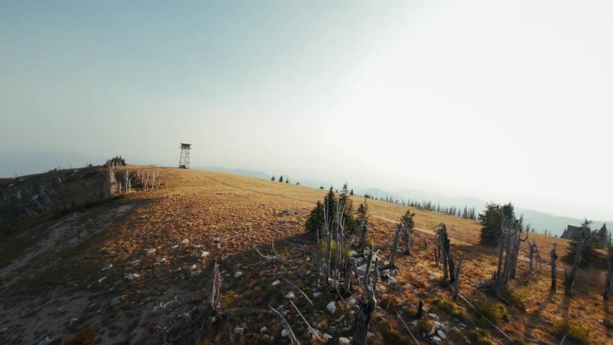 A person rides a dirt bike along a high mountain ridge toward a wooden fire lookout tower passing through a landscape of dry grass and weathered dead trees under a clear hazy sky as the camera follows the action from an aerial perspective