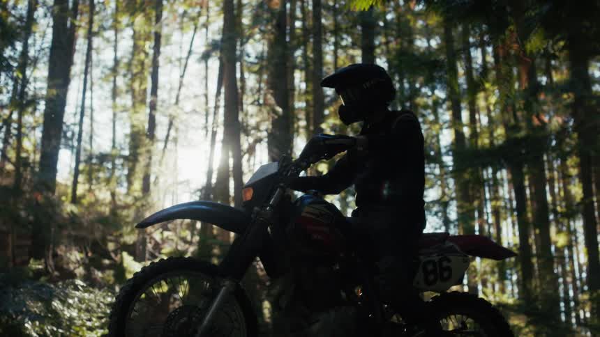 A man wearing a black helmet and dark riding gear sits on a motorcycle in a dense forest as bright sunlight shines through the tall trees behind him