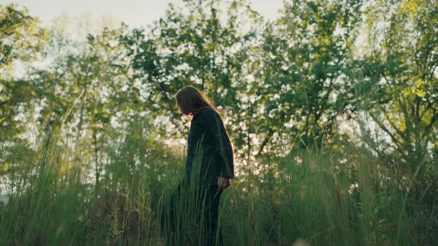 A woman wearing dark clothing walks through a field of tall grass during sunset