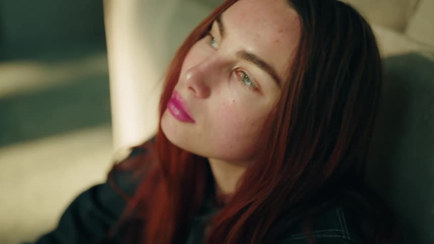 A close up shot of a young woman with long reddish brown hair and green eyes as she looks up and to the side in a sunlit room