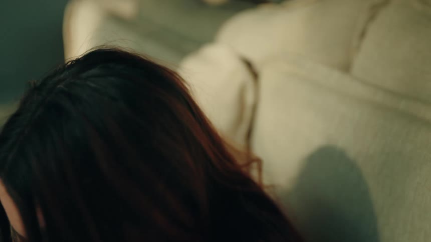 A close view of a woman with long red and brown hair leaning her head against a beige sofa in a room with warm sunlight as she looks upward and then lowers her gaze