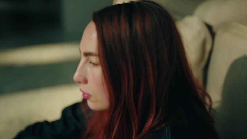 A young woman with long red hair sits on a beige sofa looking upward with a pensive expression