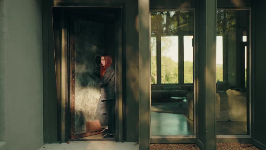 A woman with vibrant red hair is seen through large windows as she walks through a modern home then opens a heavy industrial vault door and steps outside into the afternoon light