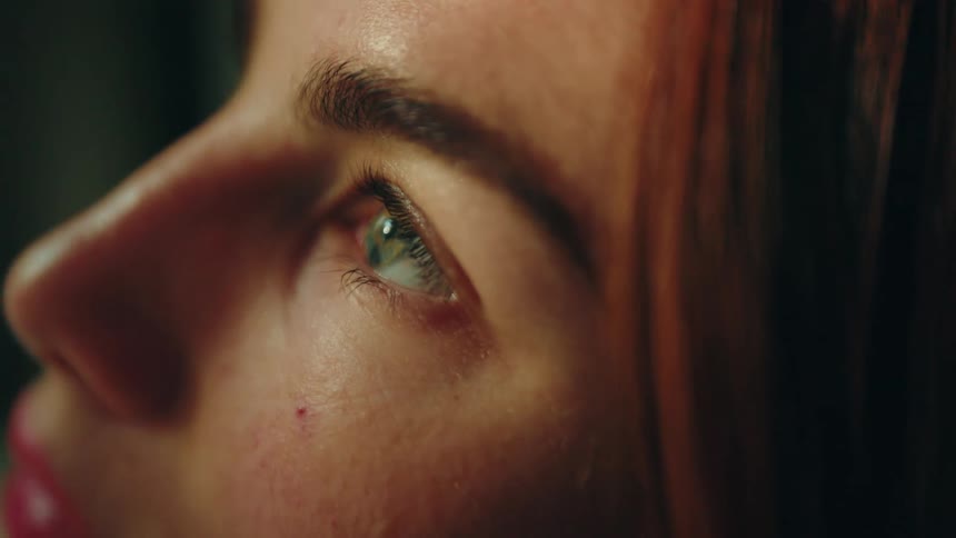 A woman with red hair looks upward in an extreme closeup and her green eye stays open reflecting light before she slowly blinks and closes it