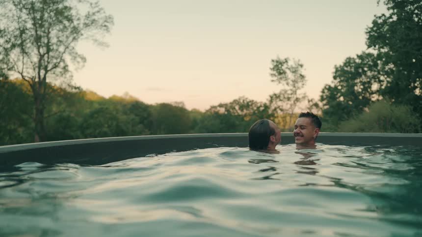 Couple in an outdoor pool exchanging looks with trees and evening sky in the background
