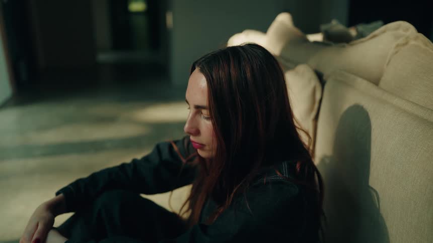A woman with long brown hair wearing a dark jacket sits on the floor leaning against a beige sofa in a dimly lit room as she looks upward with a serious expression