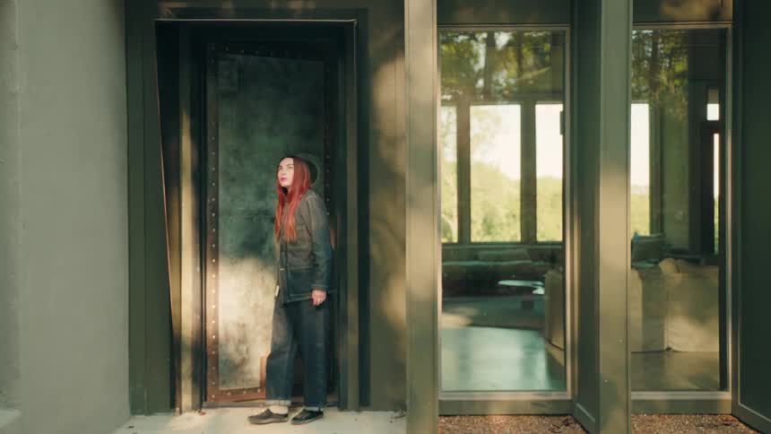 A woman with red hair walks out of a modern home through a heavy weathered metal door