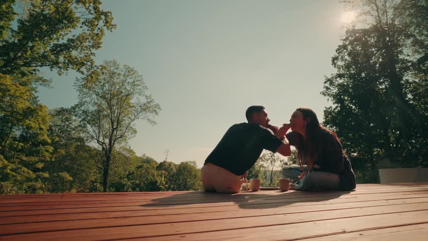 A couple sits on a sunlit wooden deck surrounded by trees feeding each other playfully as they share coffee in the warm morning light