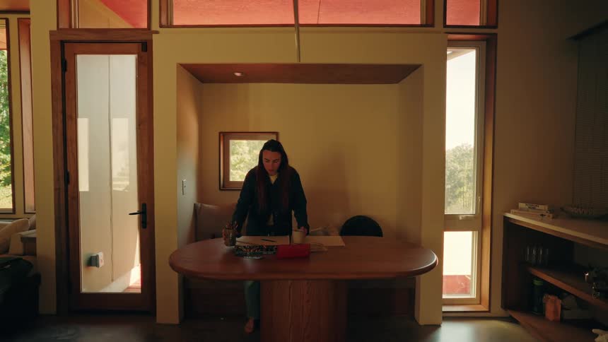 A young woman with long dark hair sits at a large wooden table in a sunlit room while wearing headphones and drinking from a white mug