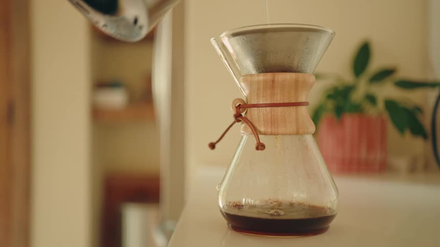 A person pours hot water from a silver kettle into a glass coffee carafe with a wood collar and paper filter as coffee drips into the bottom on a white kitchen counter
