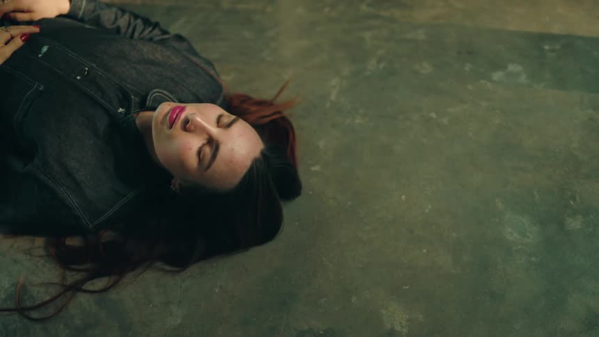 A woman lies peacefully on a concrete floor with eyes closed in a softly lit room