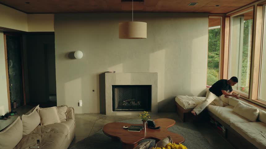 A man walks into the sunlit living room drinking and carrying a book and settles onto the cushioned bench by the window quietly sitting down to read in the calm minimalist space