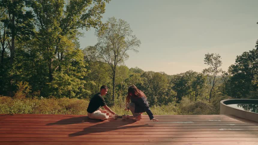 Two people sitting on a wooden deck sharing a meal outdoors with trees and greenery in the background