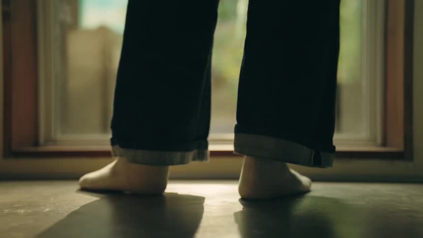 A woman wearing white socks and dark cuffed jeans walks across a sunlit room toward a large window and stop to look out at the lush green trees outside