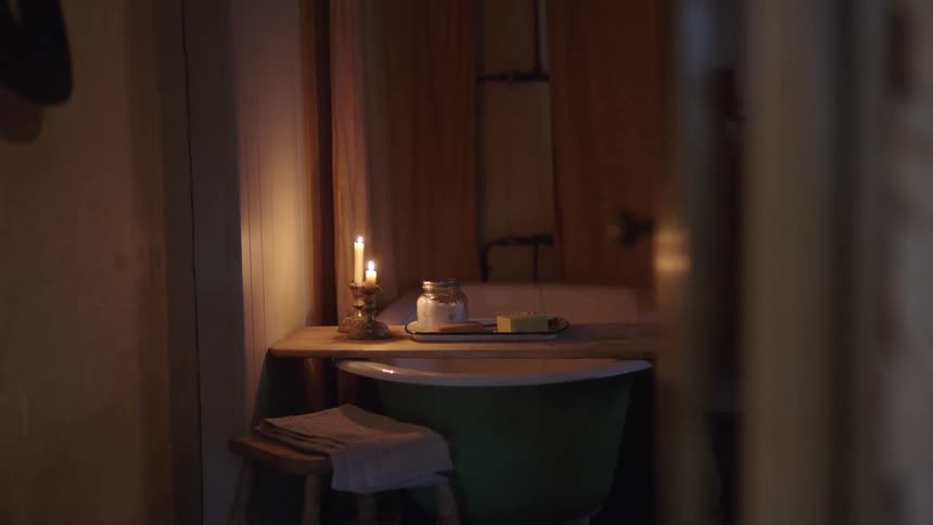 A green bathtub sits in a dimly lit bathroom with a burning candle on a wooden shelf and a small wooden stool nearby