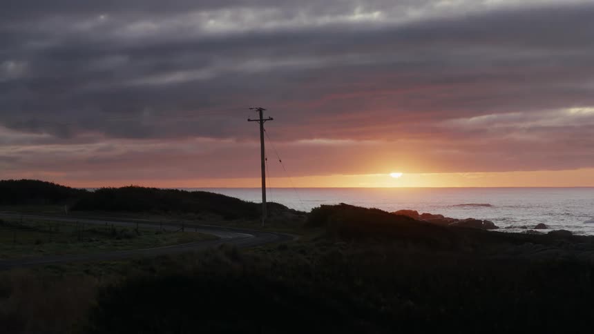 Utility pole and cables along the coast as the sun sets in a cloudy sky