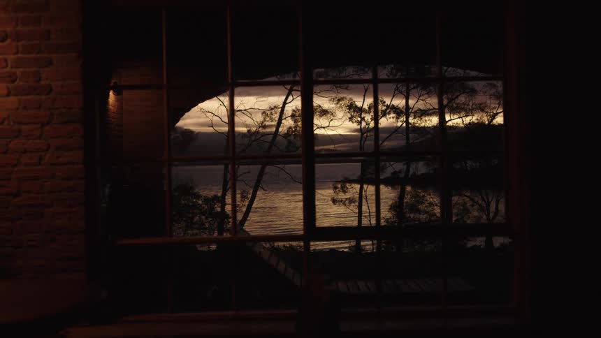 The camera looks through a large paneled window from a dark room featuring a brick wall capturing a sunset over a calm lake with silhouettes of trees and a shoreline visible against the orange sky