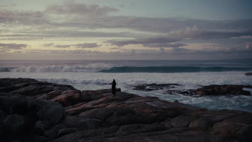 A woman in dark apparel stands still on a rocky shore watching large waves crash against the coastline under a cloudy dusk sky