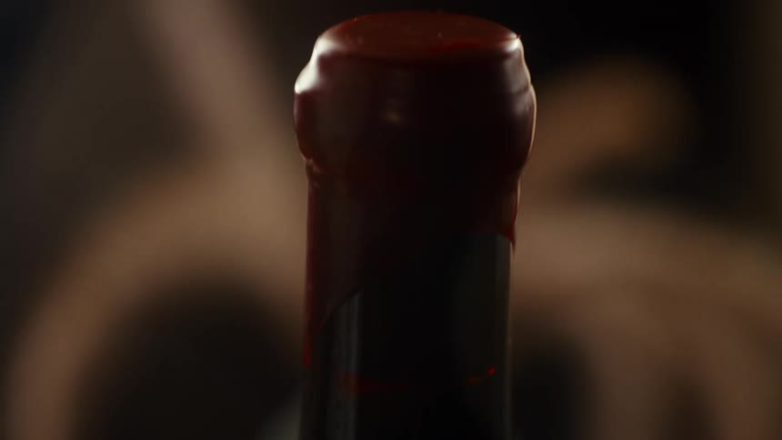 A macro camera movement travels up the neck of a dark glass wine bottle to reveal a thick red wax seal covering the top against a blurred dark background