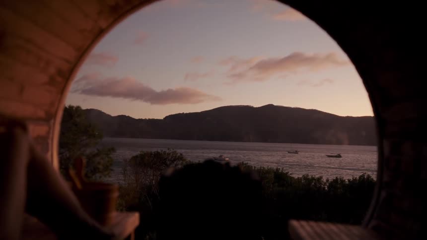 A scenic view through a round wooden window reveals a calm bay at sunset with distant hillssmall boats drift on the water while soft pink and orange clouds drift across the darkening sky