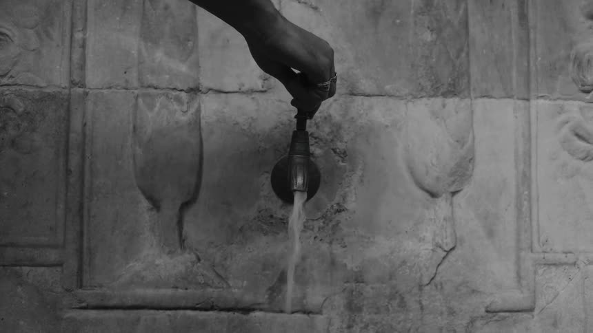 A hand reaches out and turns an ornate metal tap set into a stone wall featuring tulip carvingswater flows steadily from the spout into an unseen basin as the hand quickly moves out of the frame