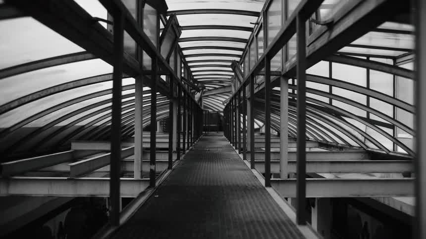A black and white view of a long empty pedestrian bridge with a curved glass roof and a metal frame