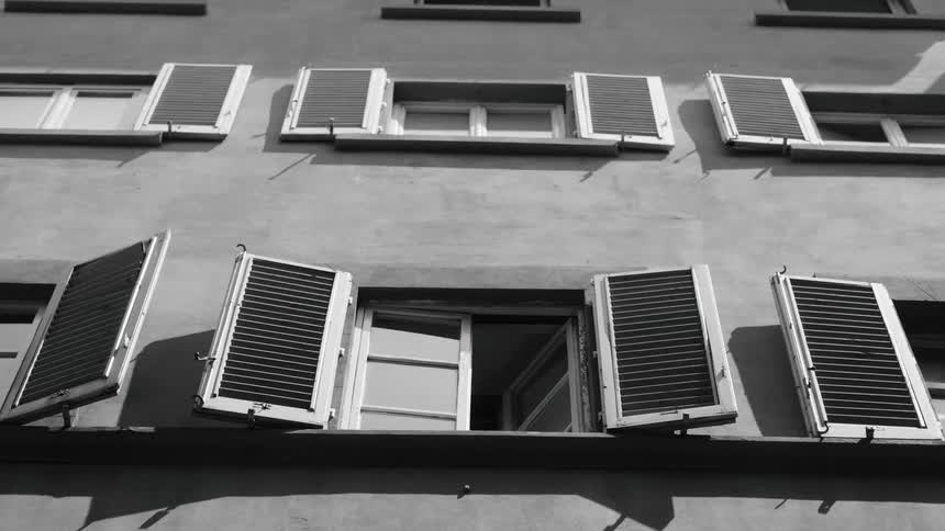 This black and white footage shows the exterior of a building featuring several windows with traditional slatted shuttersthe camera remains stationary as a single window in the lower center of the frame sits slightly open revealing a glimpse of the in