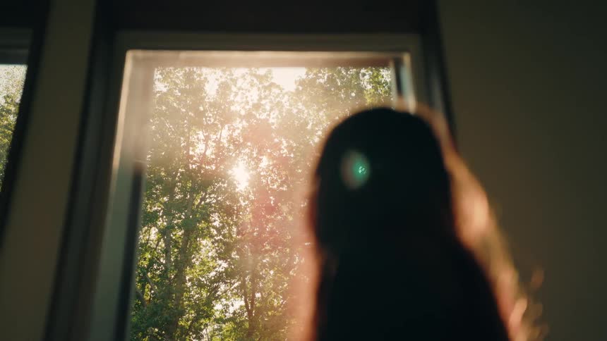 Young adult female walking to a window and looking at a view of trees and the sun before walking away into the room during day time