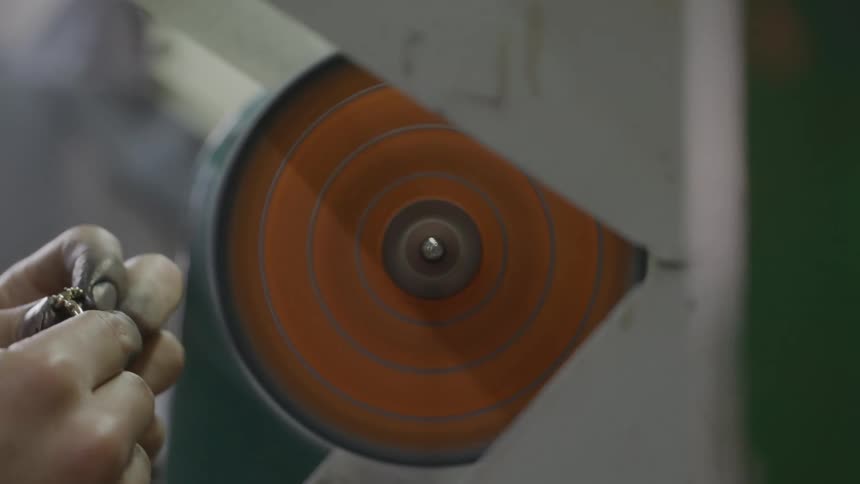 A close view of a worker using a rotating orange polishing wheel to buff a small piece of jewelry in a professional workshop